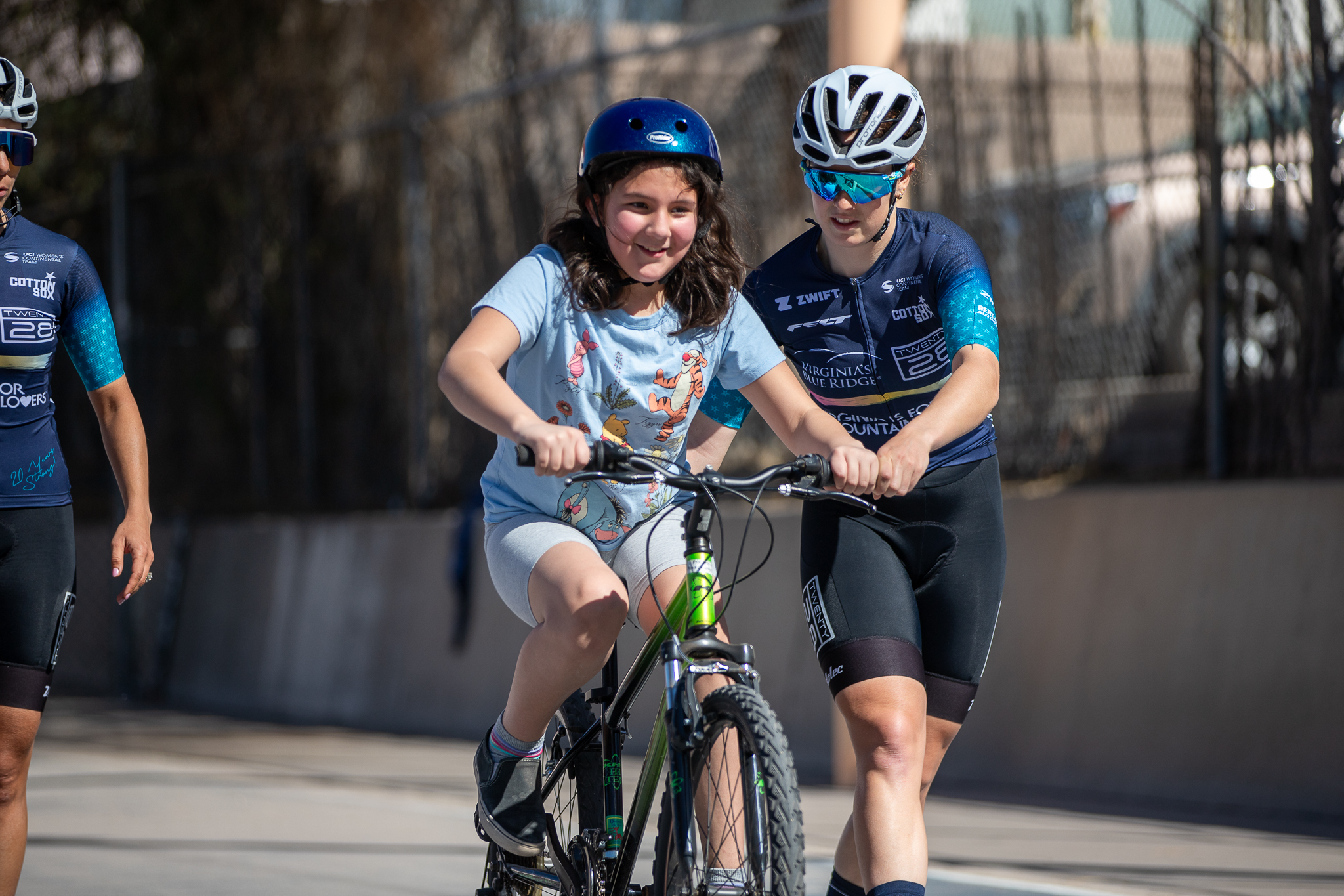 A girl rides her bike while a pro cyclist helps adjust her handlebars