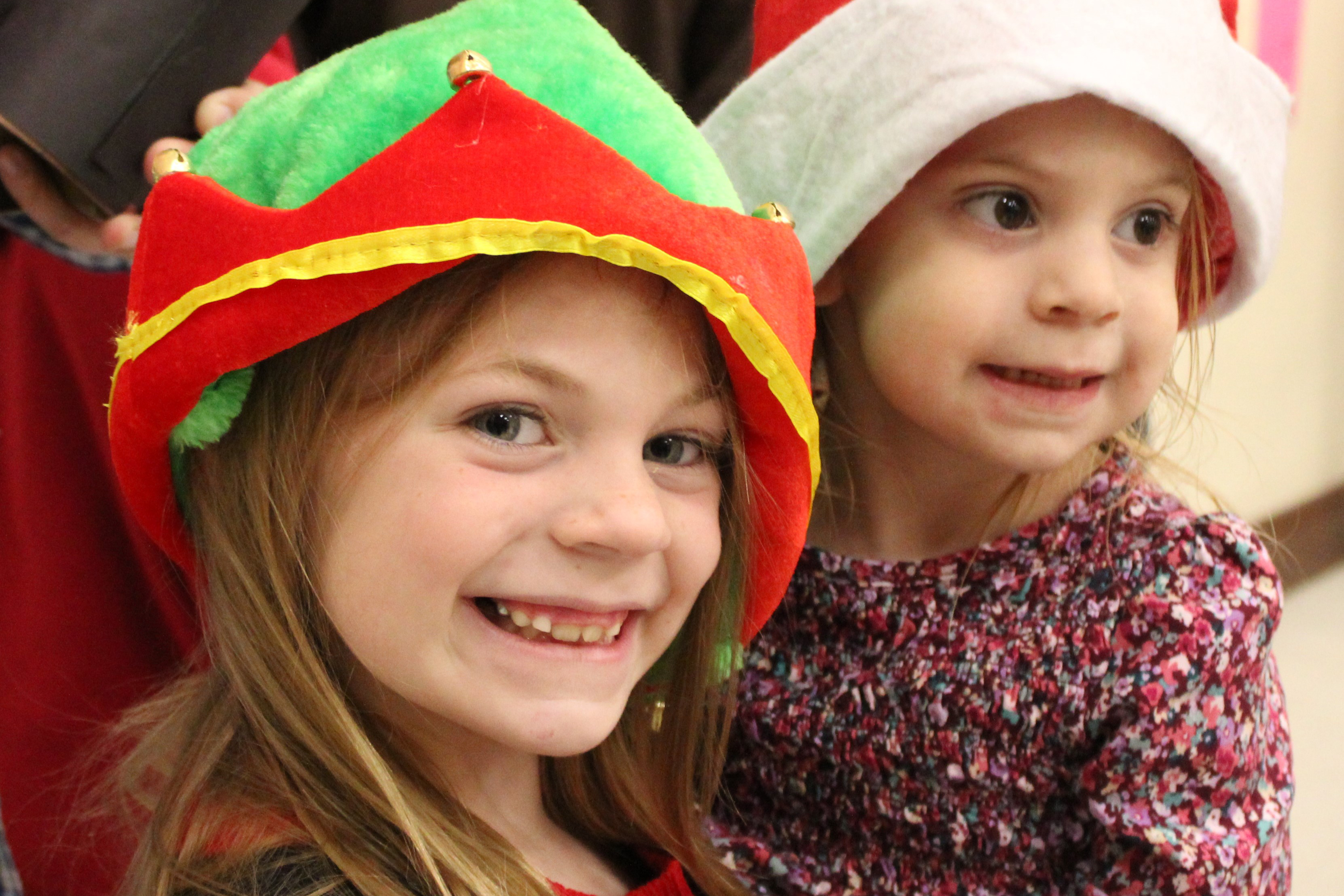 A little girl in an elf hat and a little girl in a Santa hat smile