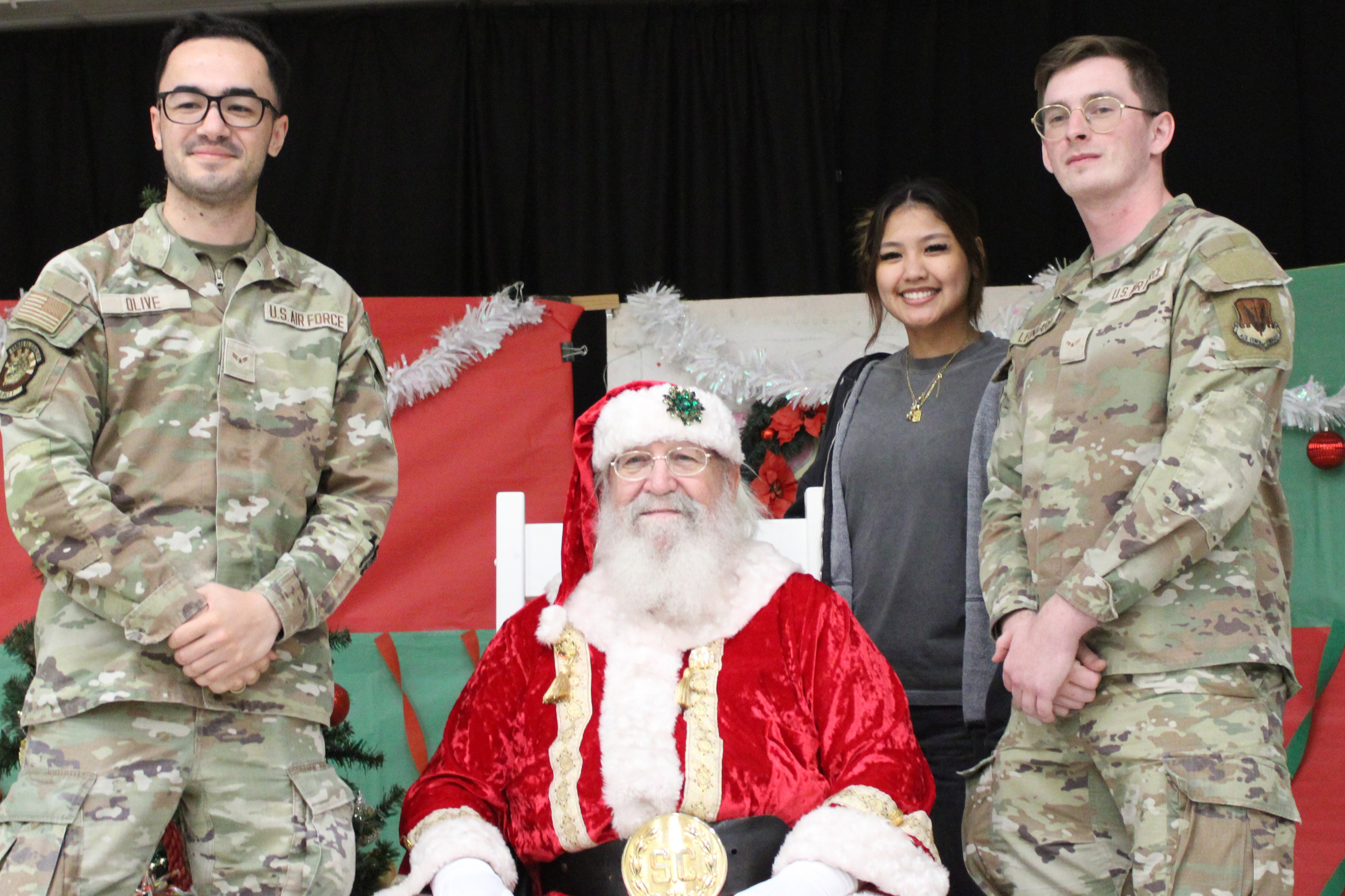 Two men in camouflage and a woman in a gray shirt pose with Santa