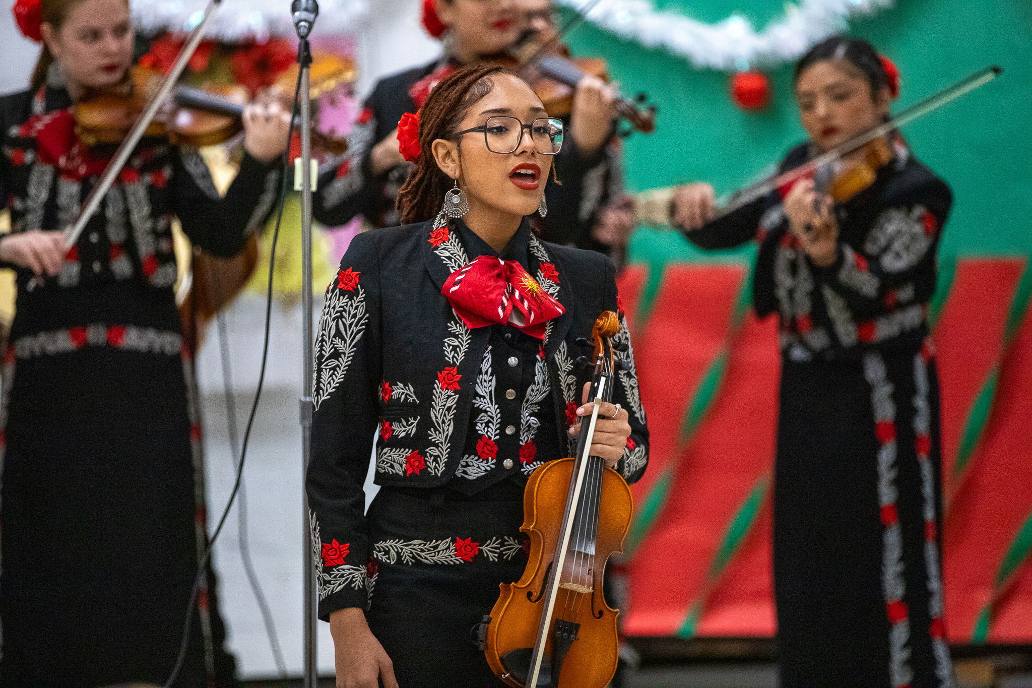 A girl in glasses holding a violin sings, while other violinists perform behind her