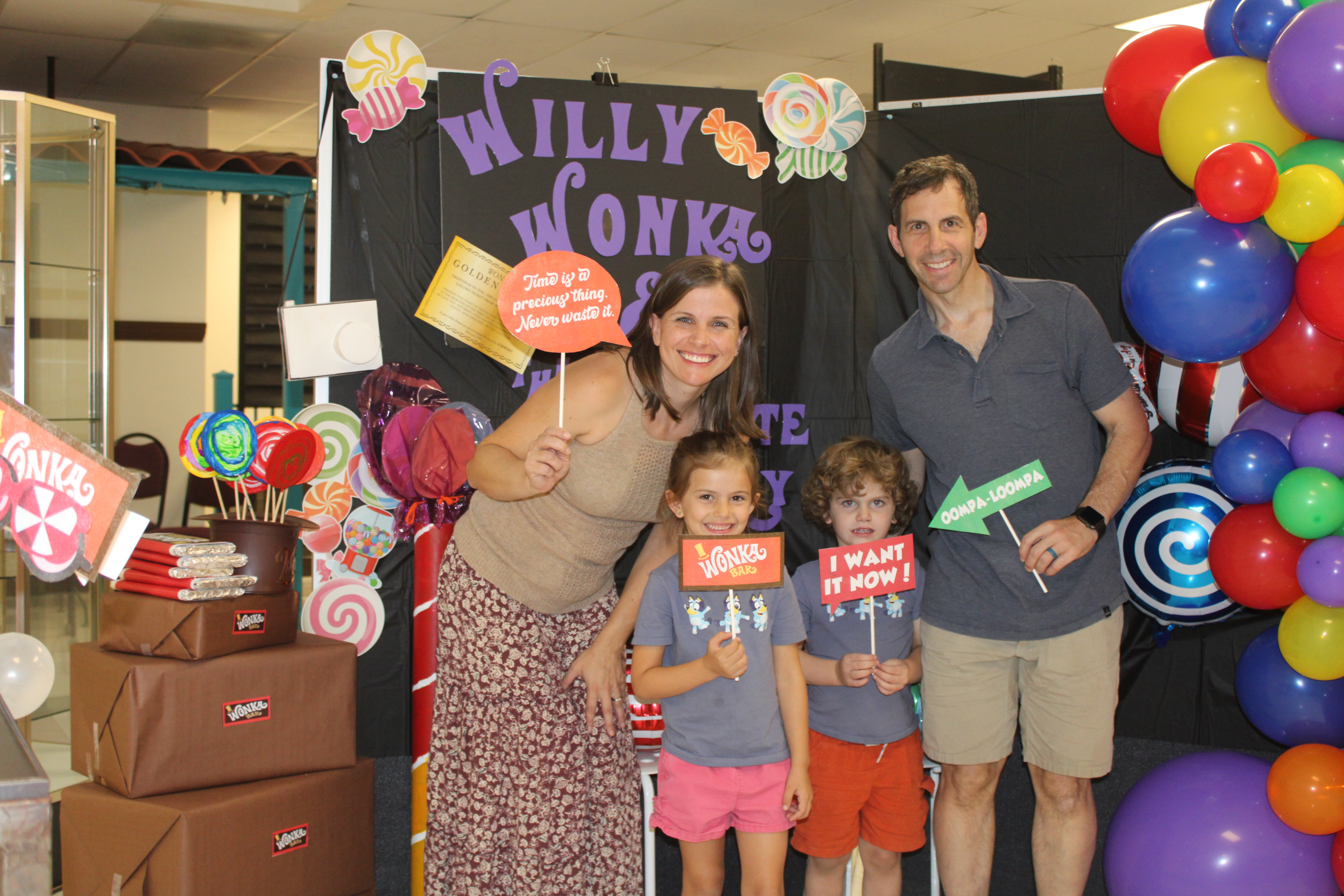 A sister and brother and their parents pose with signs in front of the Willy Wonka display