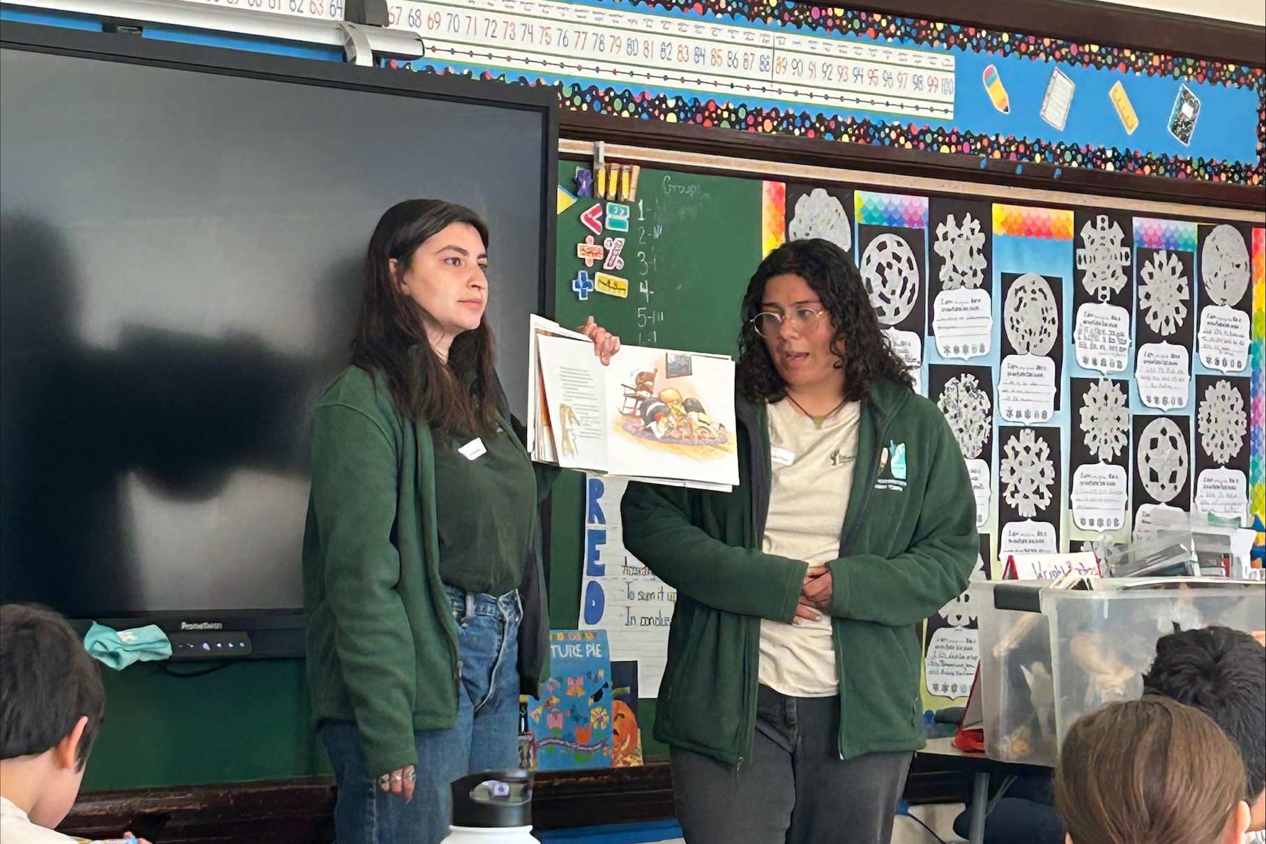 Two teachers in green stand up in front of the class to read a book