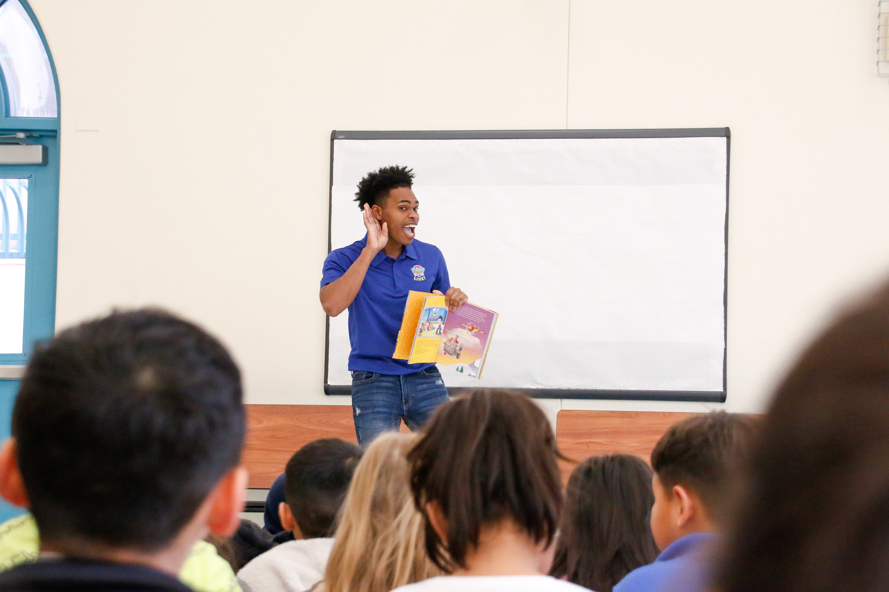 A man excitedly holds a hand up to his ear while reading a book to students