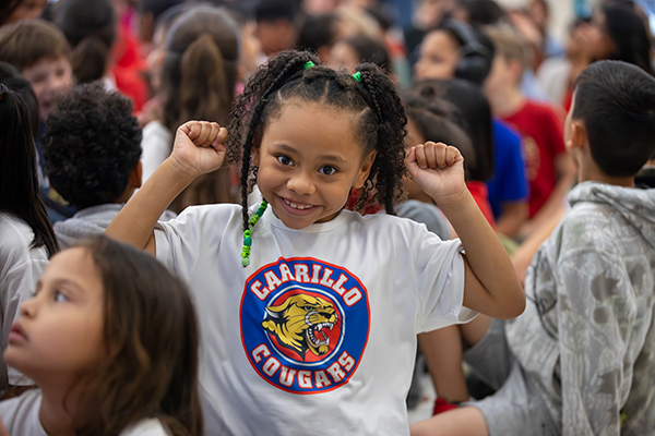 A little girl in a Carrillo Cougars shirt holds up her arms in a cheer