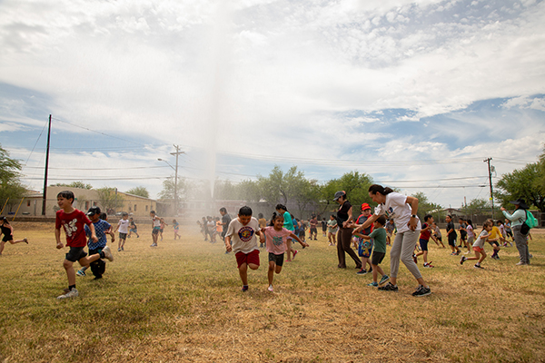Students shriek with excitement as a fire truck sprays water on them as they run across the field