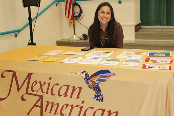 A woman smiles at a table with the Mexican American logo on it