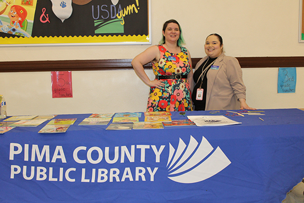 Two women smile standing behind a table with books on it and the Pima County Library logo