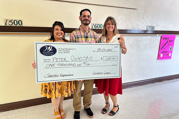 A man in a plaid shirt, center, smiles as he holds a giant check, with a woman in a yellow dress on the left and a woman in a red skirt on the right