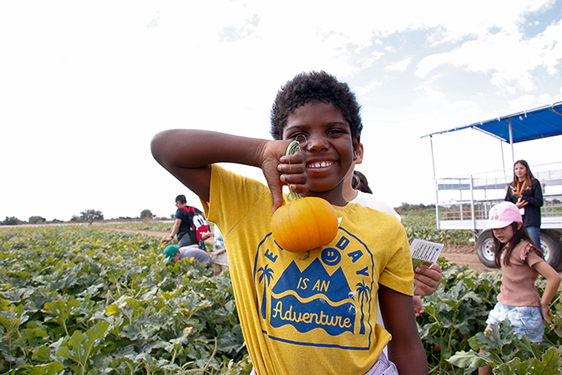 A boy in a yellow shirt smiles as he holds up a small pumpkin