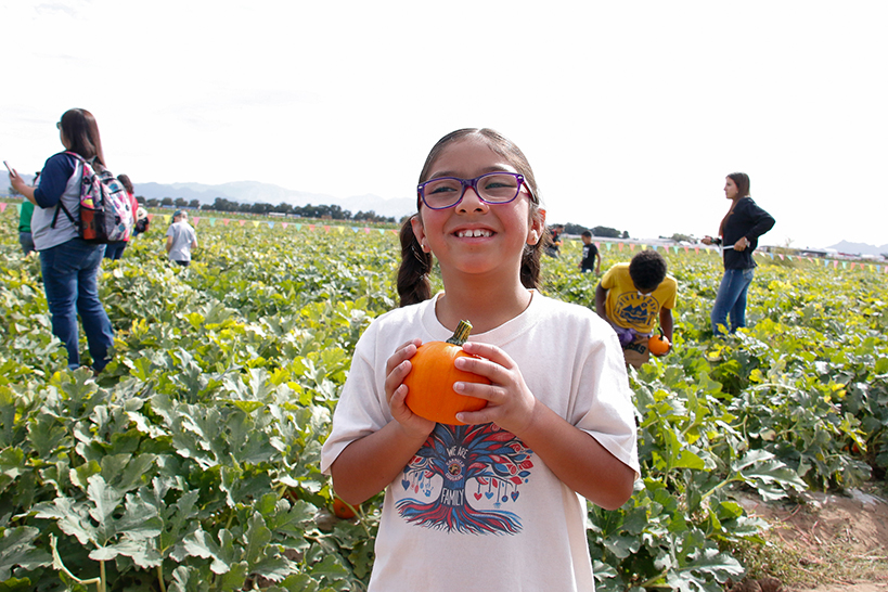 A little girl with glasses smiles as she holds up a small pumpkin