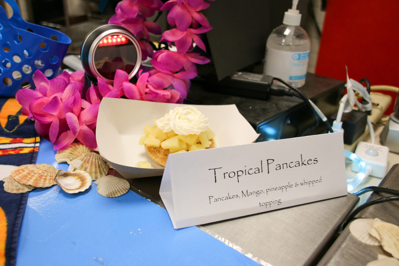 A stack of tropical pancakes layered with pineapples and whipped cream sit next to a pink lei and some seashells.