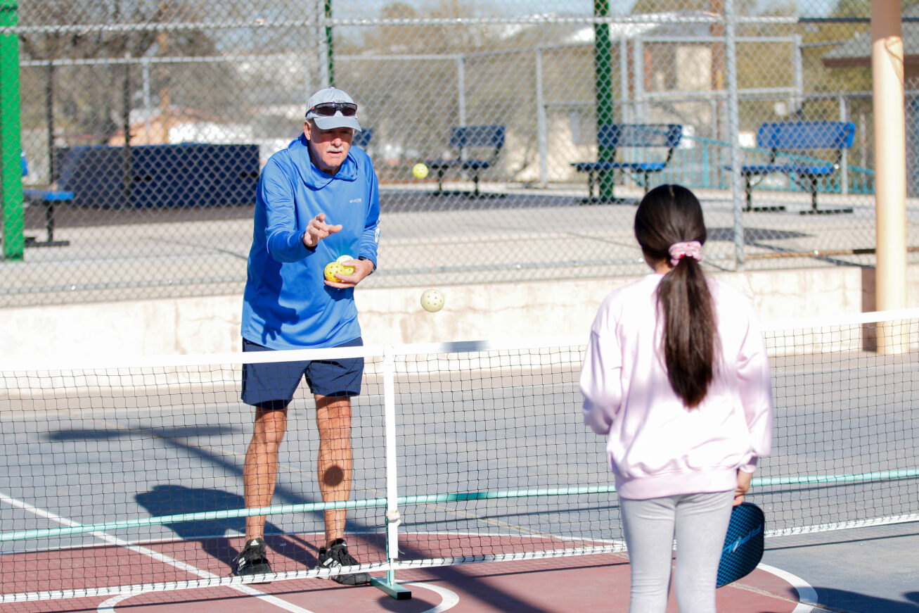 Volunteer Steve Gall demonstrates how to play pickleball while a girl in a ponytail watches his technique