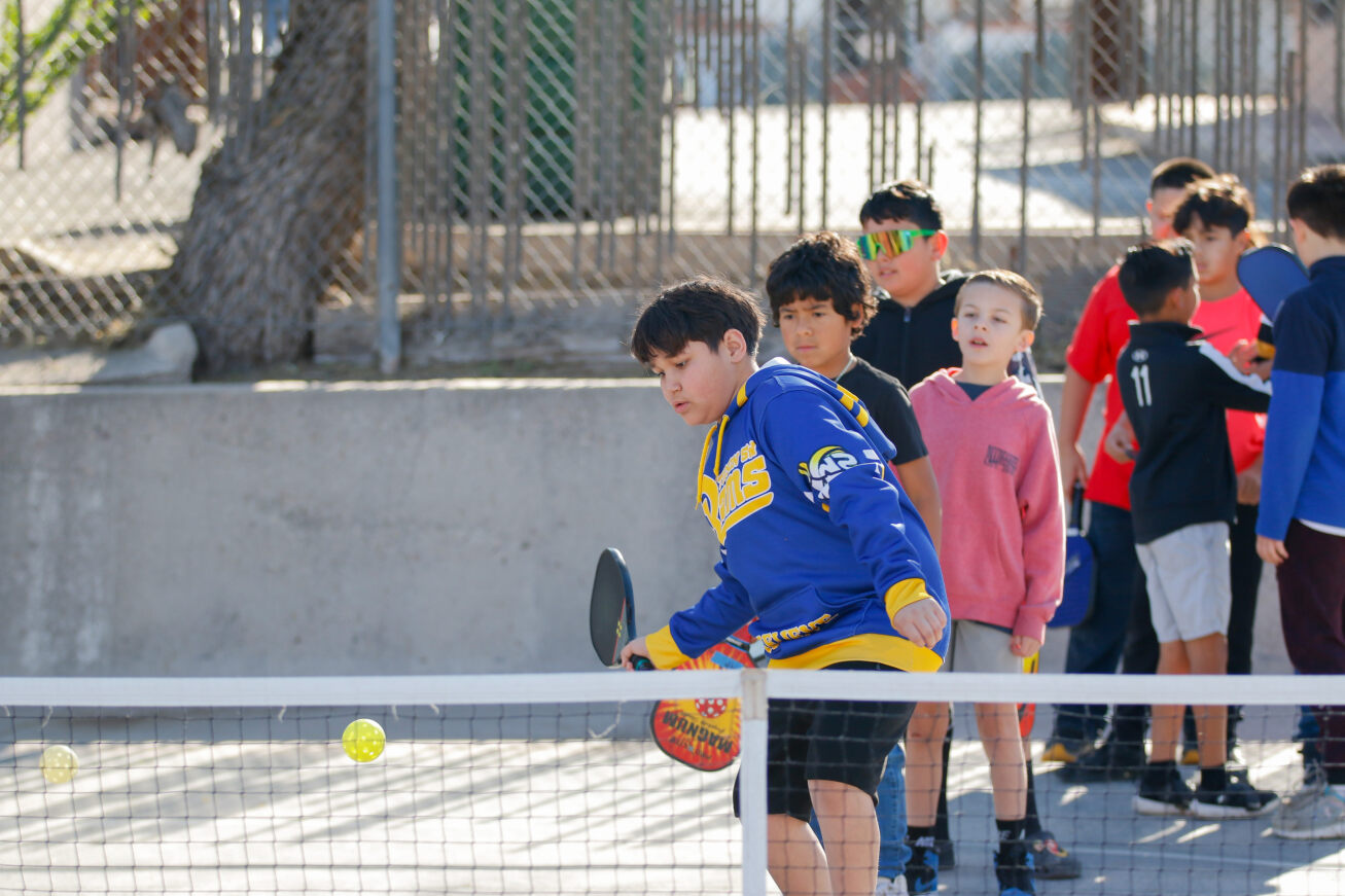 A boy in a blue hoodie hits the ball over the net while his classmates look on
