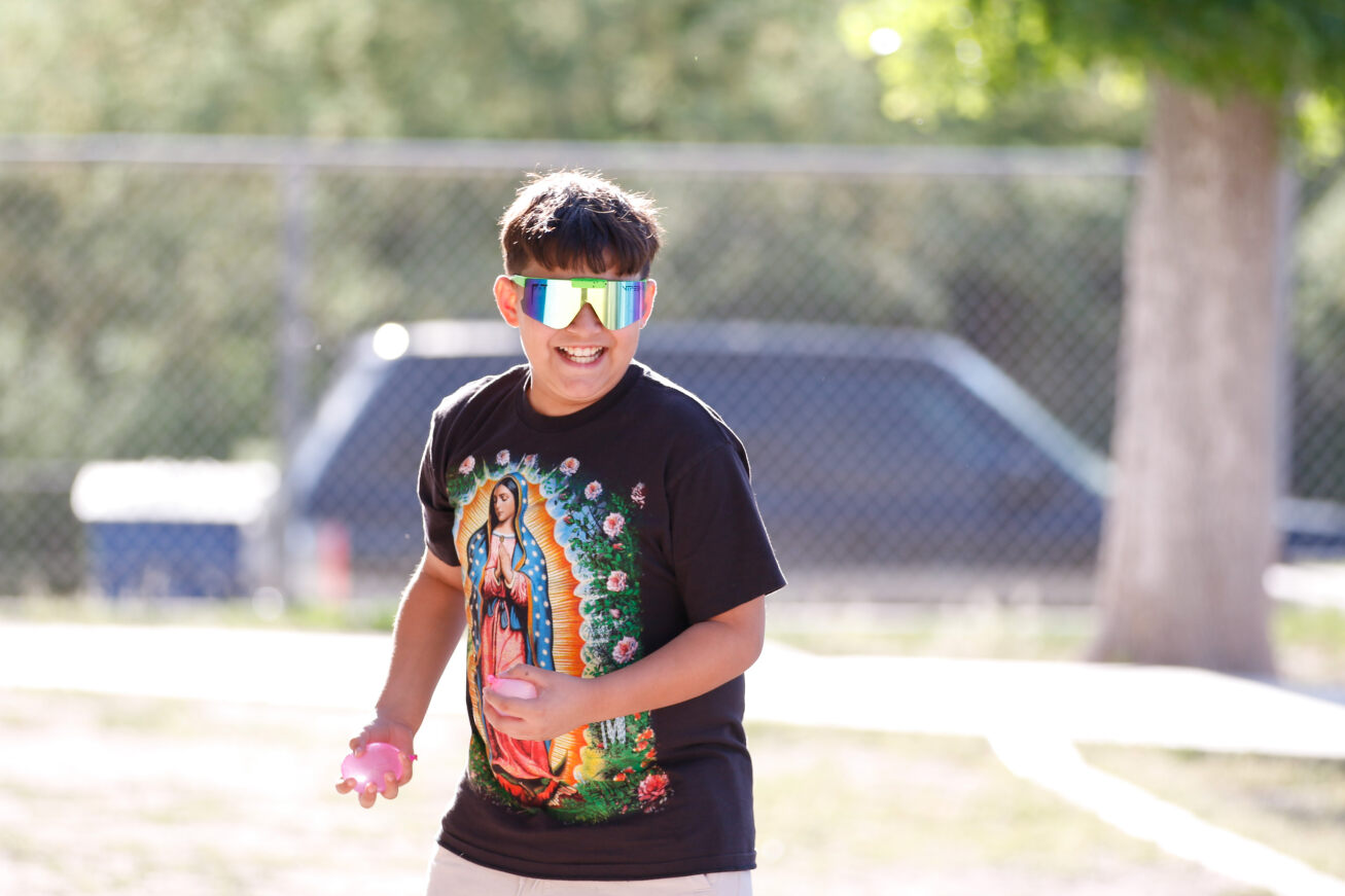A boy in sunglasses and a black shirt gets ready to throw some pink water balloons